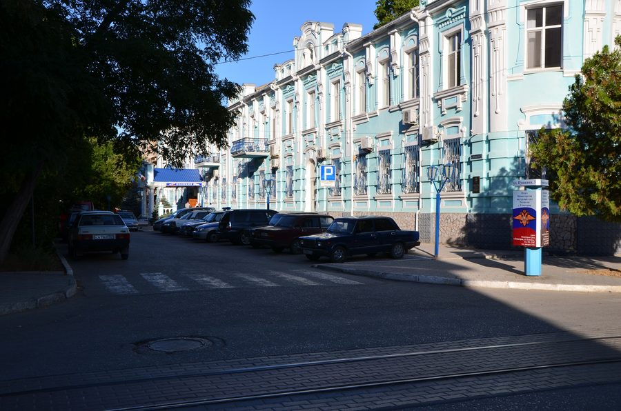 Sonnige Straße in Jewpatorija mit einem hellblauen, verzierten Gebäude, Bäumen, Autos und Straßenbahnschienen.